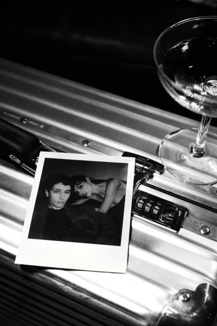 Black and white image of a photograph of two people next to a cocktail glass on a metal surface with combination lock.