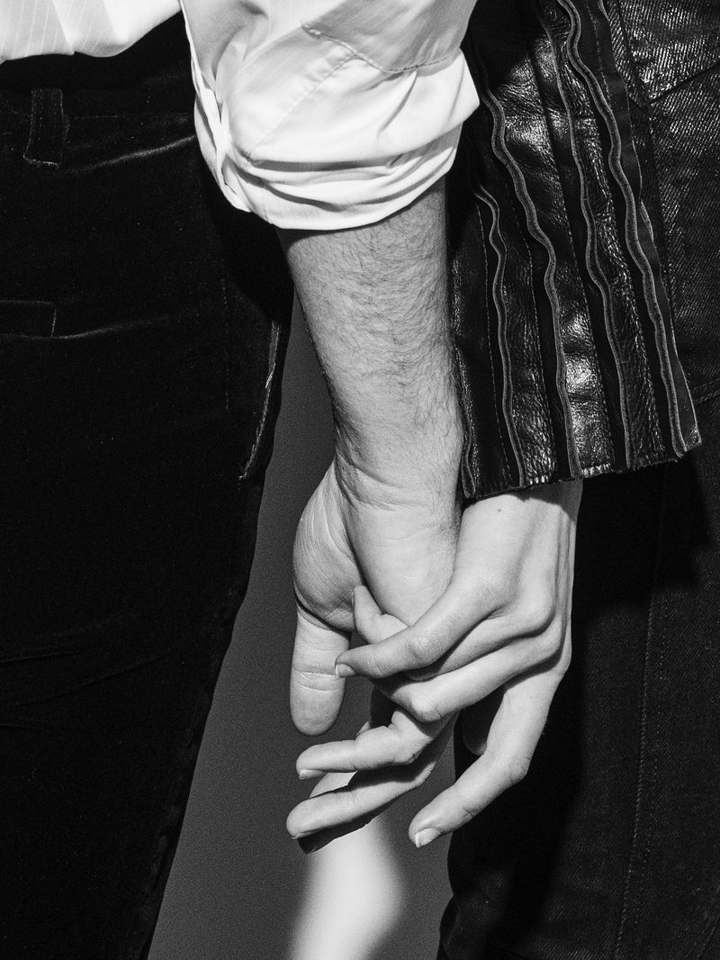 Close-up of a man and a woman holding hands, wearing dark clothing and a white button-down shirt against a neutral background.