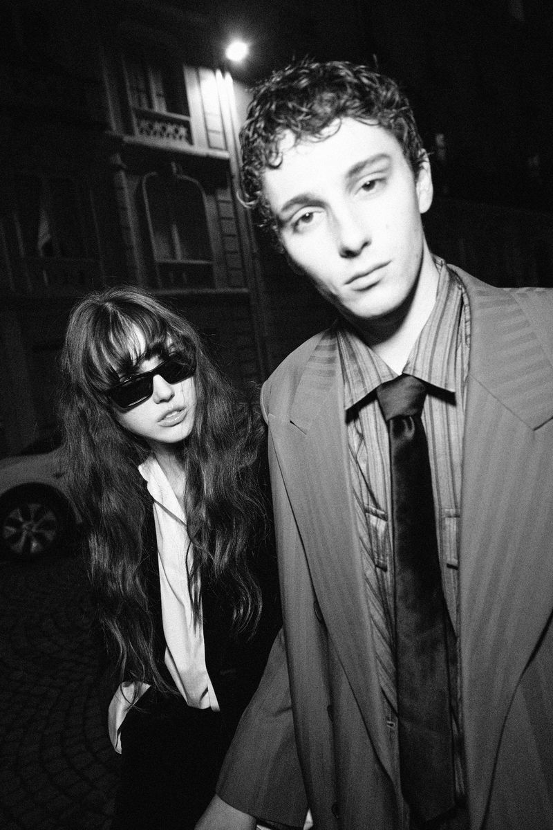 Man and woman in evening urban setting. Man wearing blazer and tie in foreground, woman with sunglasses and long hair in background.