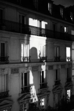 Black and white architectural photo of an apartment building at night with illuminated windows and a silhouette of a person in one window.