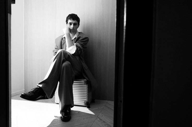 Black and white portrait of a stylish man sitting on luggage, wearing a suit and black shoes, with an interior background.