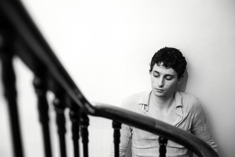 Monochrome portrait of a young adult man with curly hair, wearing a casual shirt, seated indoors near a dark wooden staircase.