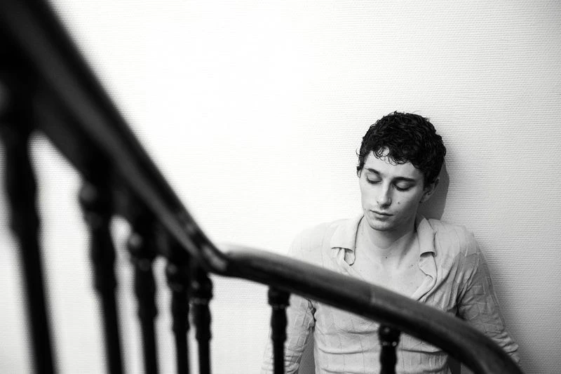 Monochrome portrait of a young adult man with curly hair, wearing a casual shirt, seated indoors near a dark wooden staircase.