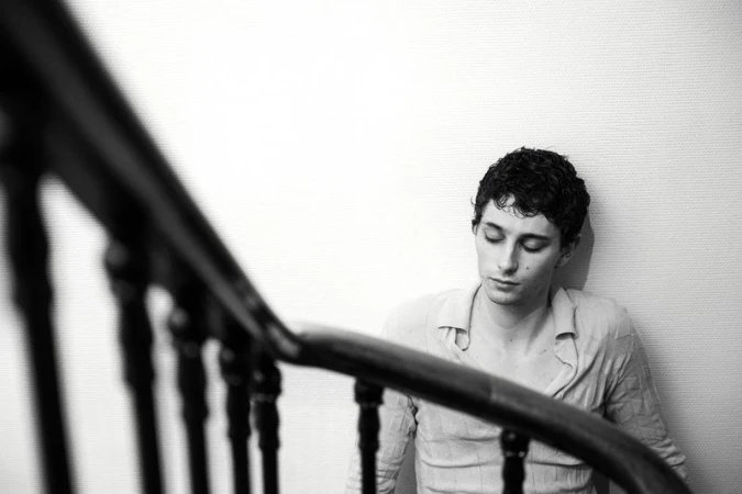 Monochrome portrait of a young adult man with curly hair, wearing a casual shirt, seated indoors near a dark wooden staircase.