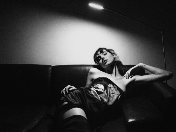 Monochrome fashion editorial image featuring a female model reclining on a dark leather sofa beneath a floor lamp