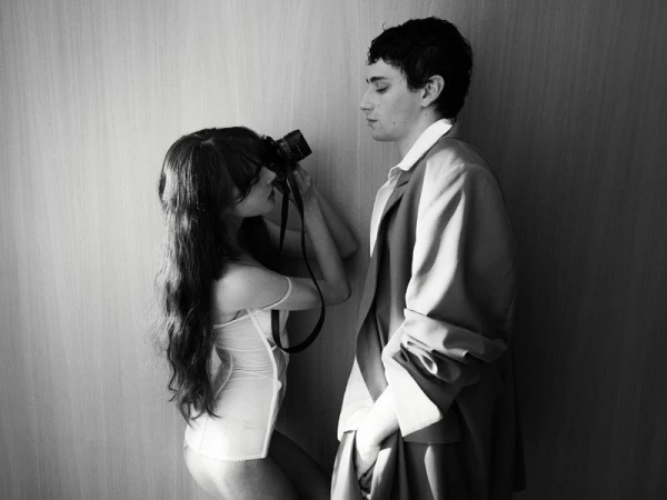 A young brunette woman in a white top takes a photo of a slender young man in a blazer against a neutral-toned wall, captured in black and white.