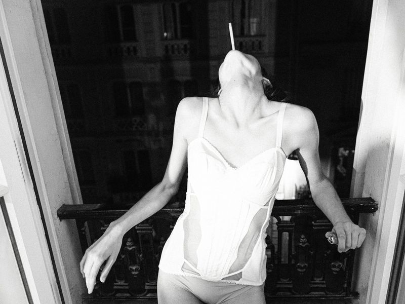 Monochrome portrait of a slender adult female in underwear and a corset, holding a cigarette in her mouth and leaning on a balcony.