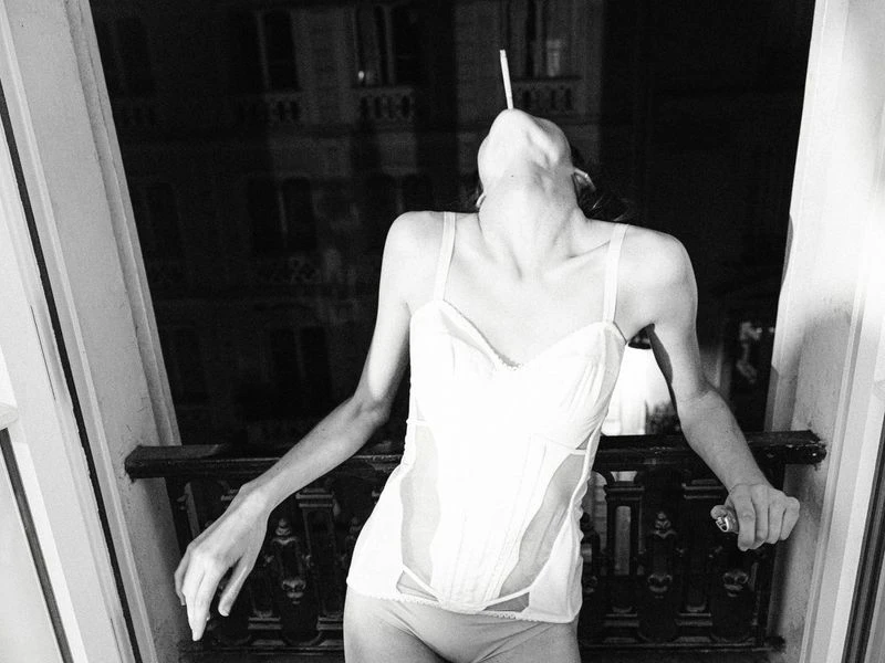 Monochrome portrait of a slender adult female in underwear and a corset, holding a cigarette in her mouth and leaning on a balcony.