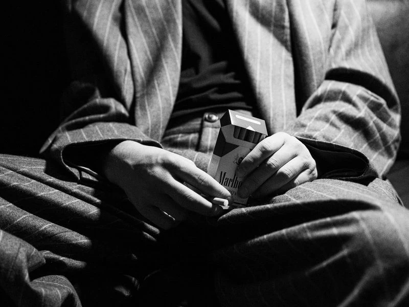 Close-up black and white shot of a person holding a cigarette and a Marlboro cigarette pack while wearing a striped suit.