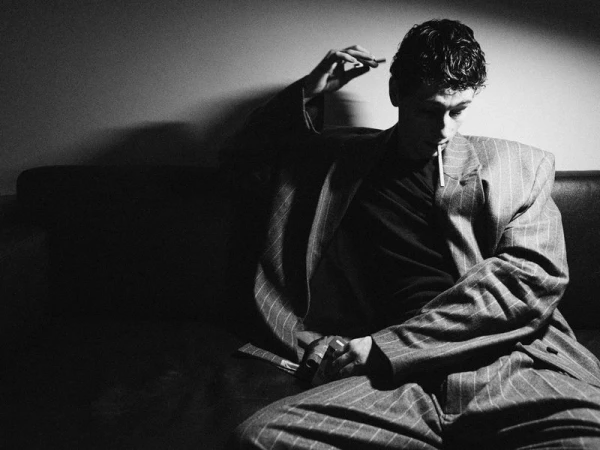 Young man in a pinstriped suit sitting on a dark couch with a cigarette in his mouth, illuminated by high-contrast studio lighting.