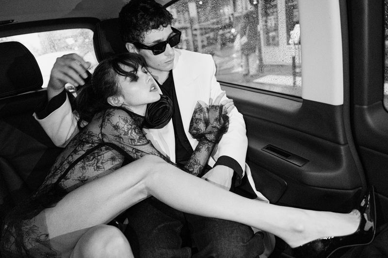 Black and white fashion shot in the back of a car features a man in a white suit embracing a woman in a lace dress, creating an intimate mood.