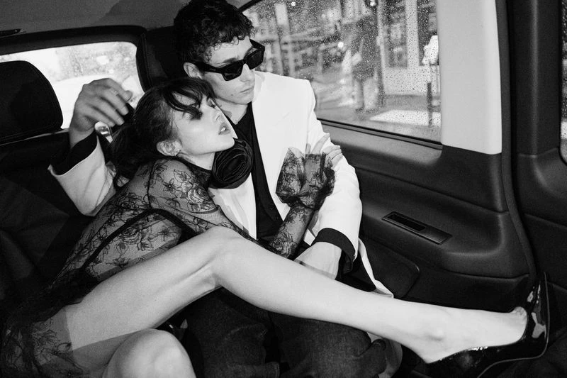 Black and white fashion shot in the back of a car features a man in a white suit embracing a woman in a lace dress, creating an intimate mood.