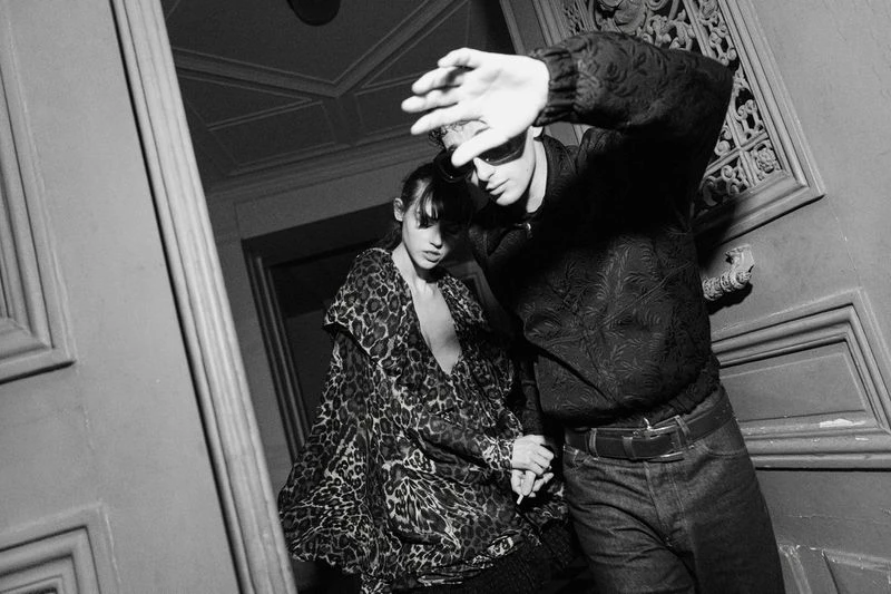 Black and white fashion shot of a stylish couple walking down stairs; the man shields his face, and the woman wears a leopard print coat.