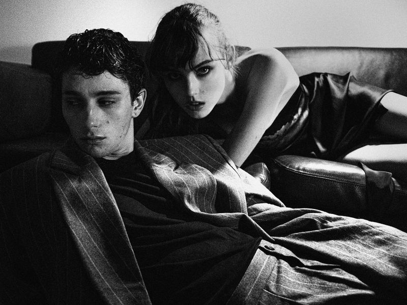 A young man in a pinstripe blazer and woman in a sleeveless top lounging on a dark sofa in a high-contrast black and white photograph.