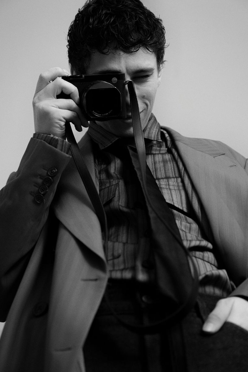Monochrome studio portrait of a man taking a photo with a camera, wearing a blazer, patterned shirt, and tie in a minimalist setting.