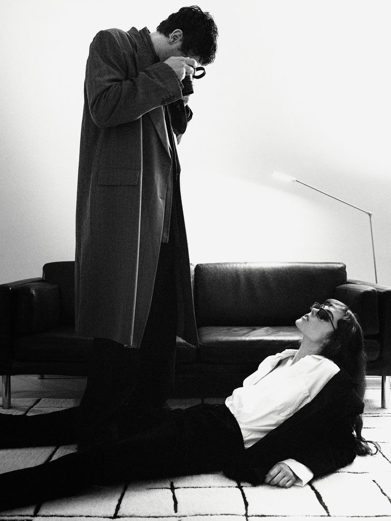 Monochrome image of a man in a coat taking a photo of a woman in a suit lying on the floor, near a black couch in an indoor setting.