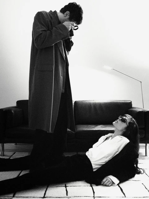 Monochrome image of a man in a coat taking a photo of a woman in a suit lying on the floor, near a black couch in an indoor setting.