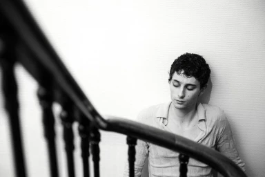 Monochrome portrait of a young man seated against a wall next to a dark wooden staircase railing.