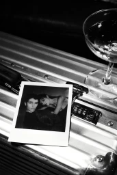 Black and white editorial image showing a polaroid photograph of a couple and a cocktail glass resting on a metallic suitcase