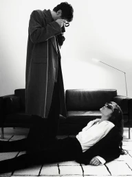 A man in a long coat taking a photo while standing over a woman lying on a patterned rug in front of a leather sofa.