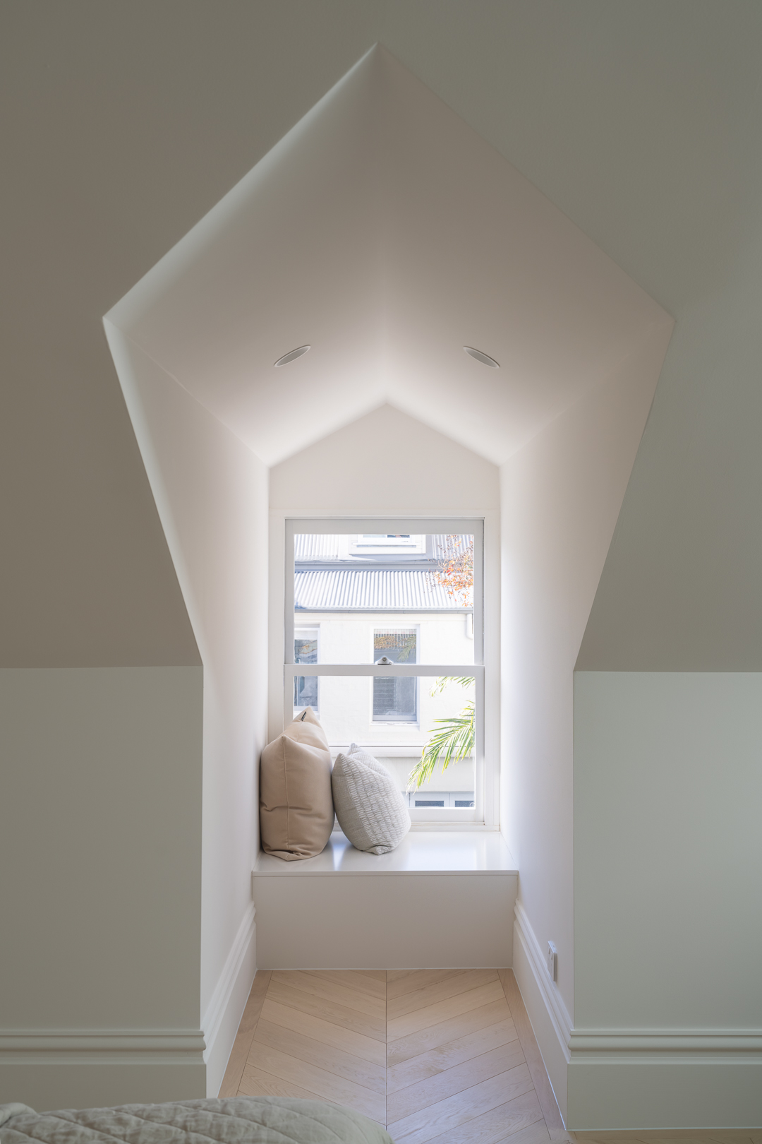 Indoor architectural shot of a white-walled window nook featuring cushions, bright natural light, and herringbone wood flooring.