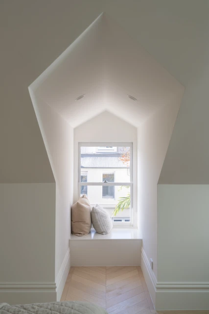 Indoor architectural shot of a white-walled window nook featuring cushions, bright natural light, and herringbone wood flooring.