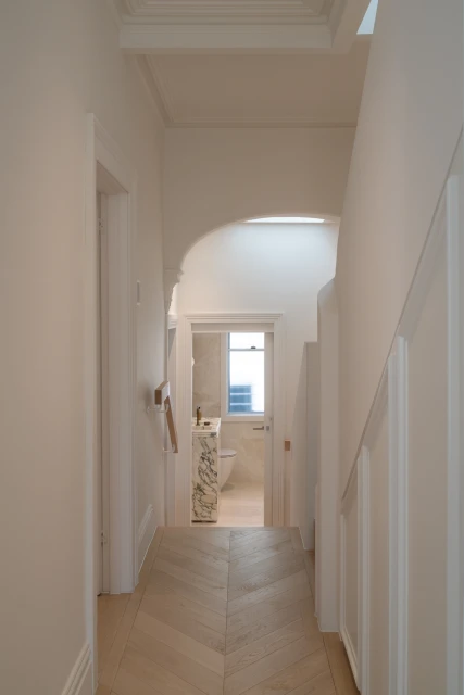 Hallway interior with herringbone wood floors, white walls, and an open doorway leading to a bathroom with natural light.