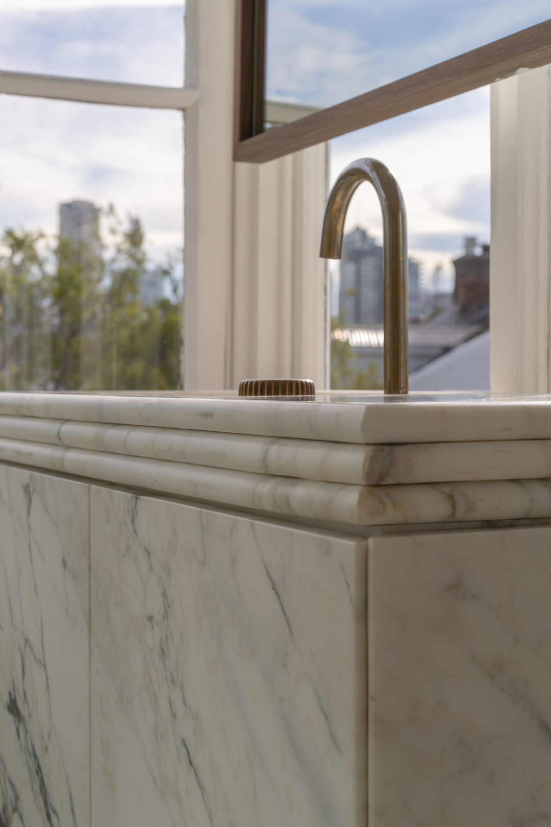Close-up on a marble countertop with a brass faucet and running water, with a cityscape visible through a nearby window.