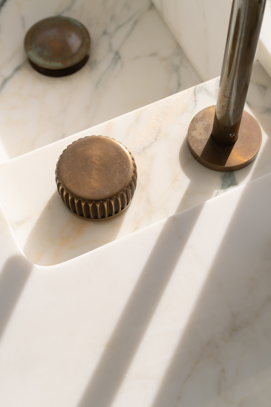 Close-up of bronze faucet knobs and fixture on a marble sink with veining detail and linear shadows creating abstract patterns.
