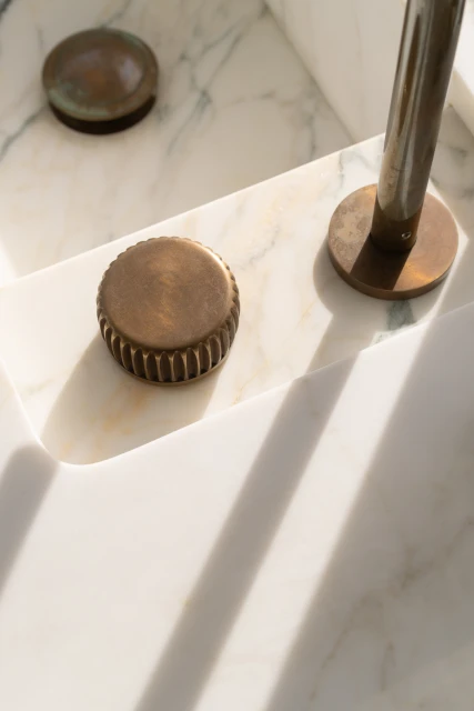 Close-up of bronze faucet knobs and fixture on a marble sink with veining detail and linear shadows creating abstract patterns.