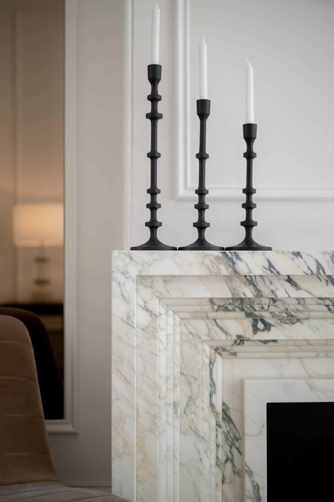 Three black candlestick holders with white candles are displayed on a marble fireplace mantel in a bright, modern home interior.