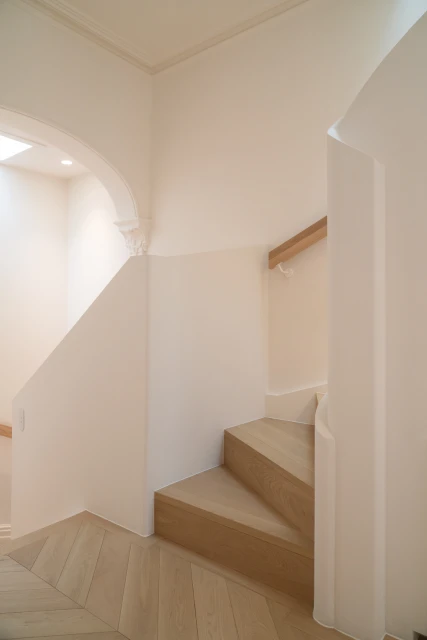 Indoor staircase with light wood steps and herringbone pattern wood flooring, white walls, arched doorway, and natural light from above.