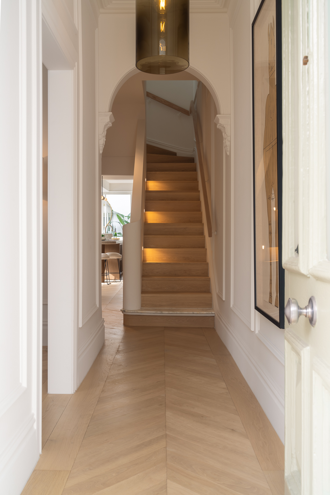 Hallway with herringbone wood floor, stairs leading up, and a doorway leading into a kitchen area in a modern, bright house.