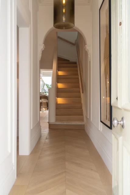 Hallway with herringbone wood floor, stairs leading up, and a doorway leading into a kitchen area in a modern, bright house.