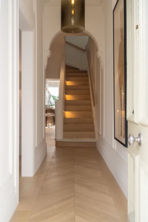 Hallway with herringbone wood floor, stairs leading up, and a doorway leading into a kitchen area in a modern, bright house.