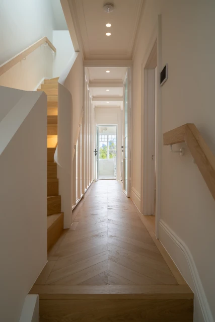 Hallway view featuring a wooden staircase and herringbone pattern flooring that leads to a white-framed glass door at the end of the corridor.