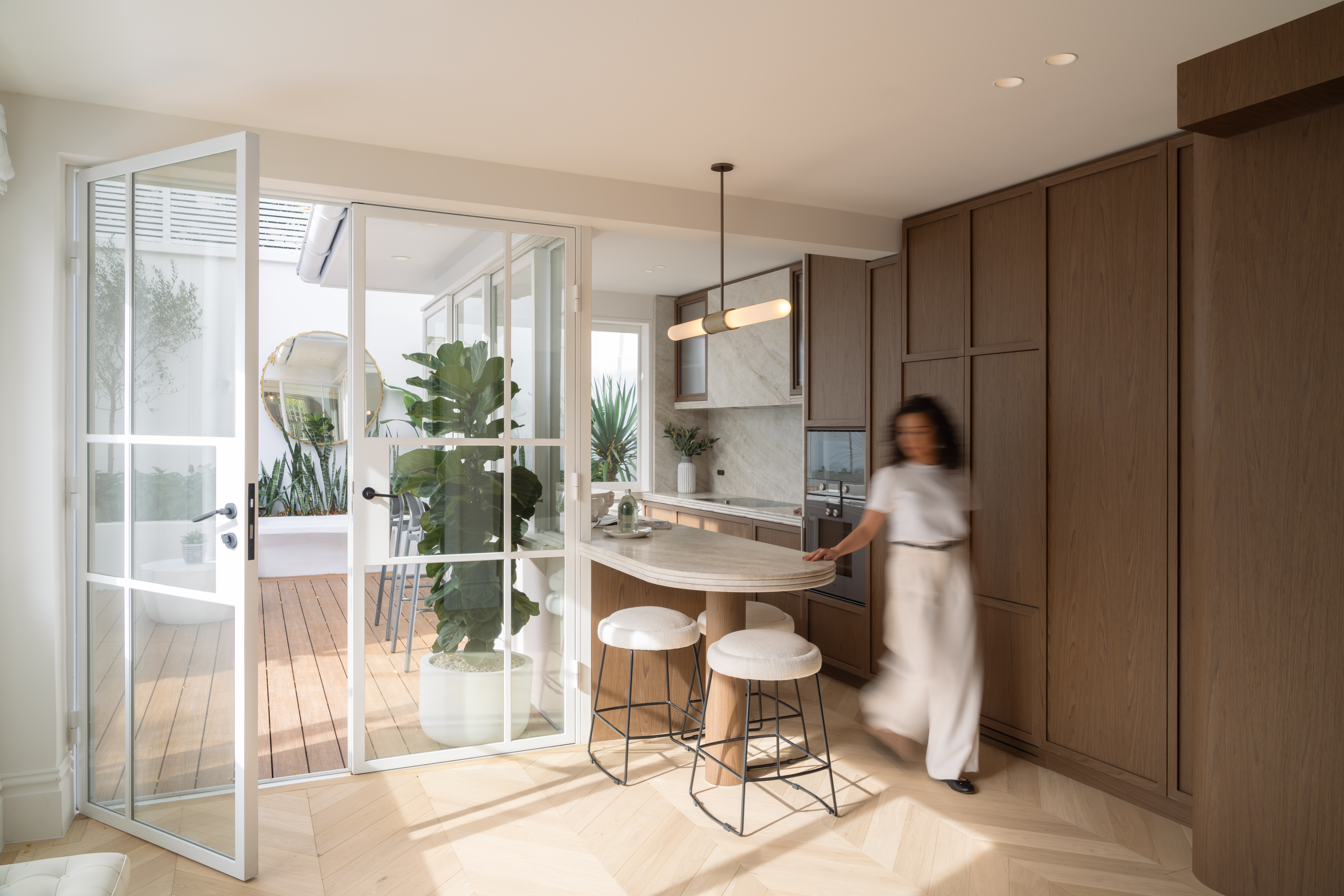 Modern kitchen interior with an island, stools, cabinetry, and open doors leading to an outdoor patio with plants and furniture.