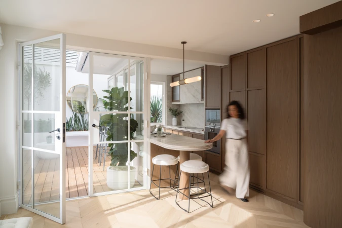 Modern kitchen interior with an island, stools, cabinetry, and open doors leading to an outdoor patio with plants and furniture.