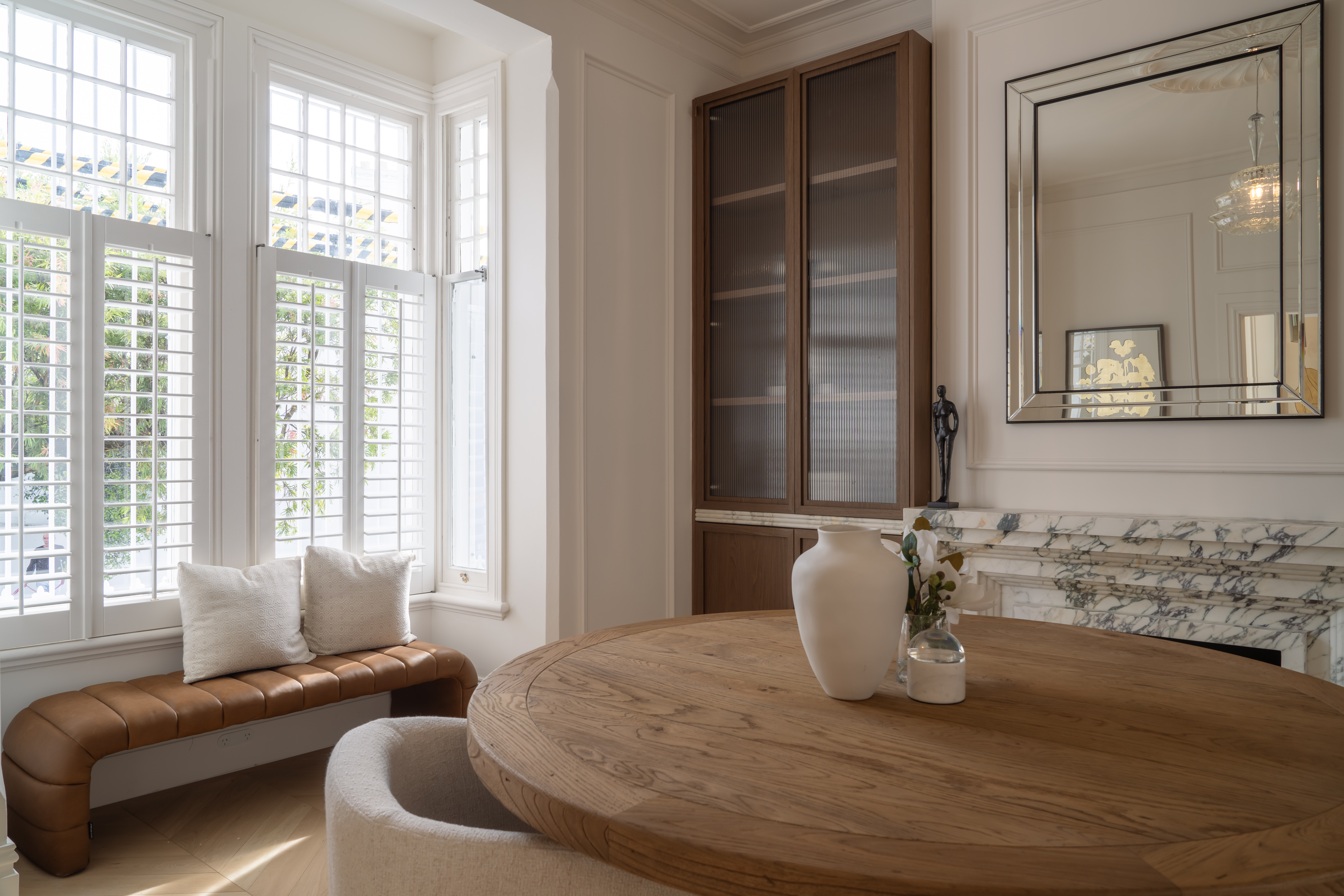 Interior design with round wooden table, upholstered bench, fireplace, and window with shutters in a brightly lit living space.
