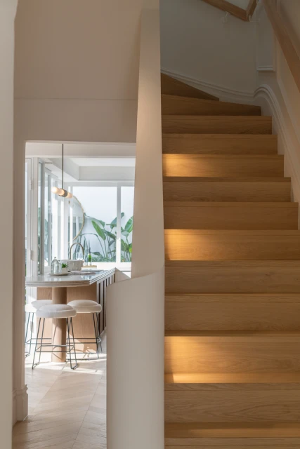 Interior view of a modern home with light wood stairs ascending, a glimpse into a sunlit kitchen with white stools and plants.