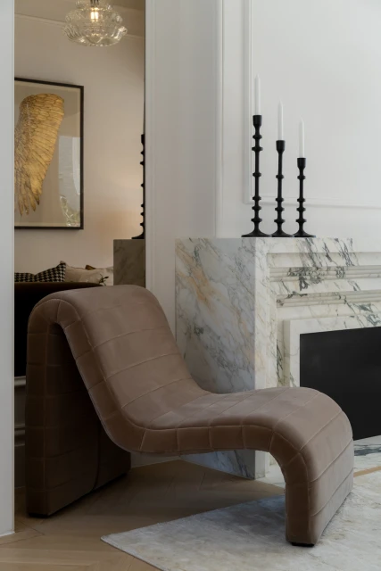 Interior with a modern chaise lounge in the foreground, marble fireplace, and a decorative painting creating an elegant, neutral-toned space.