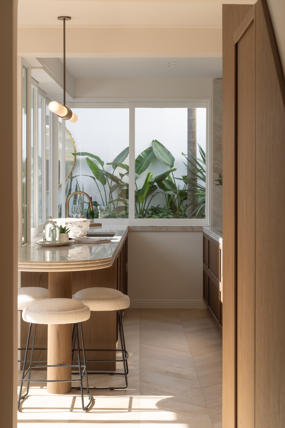 Indoor kitchen area with a marble countertop, wooden stools, and a view to the outside garden through a large window.