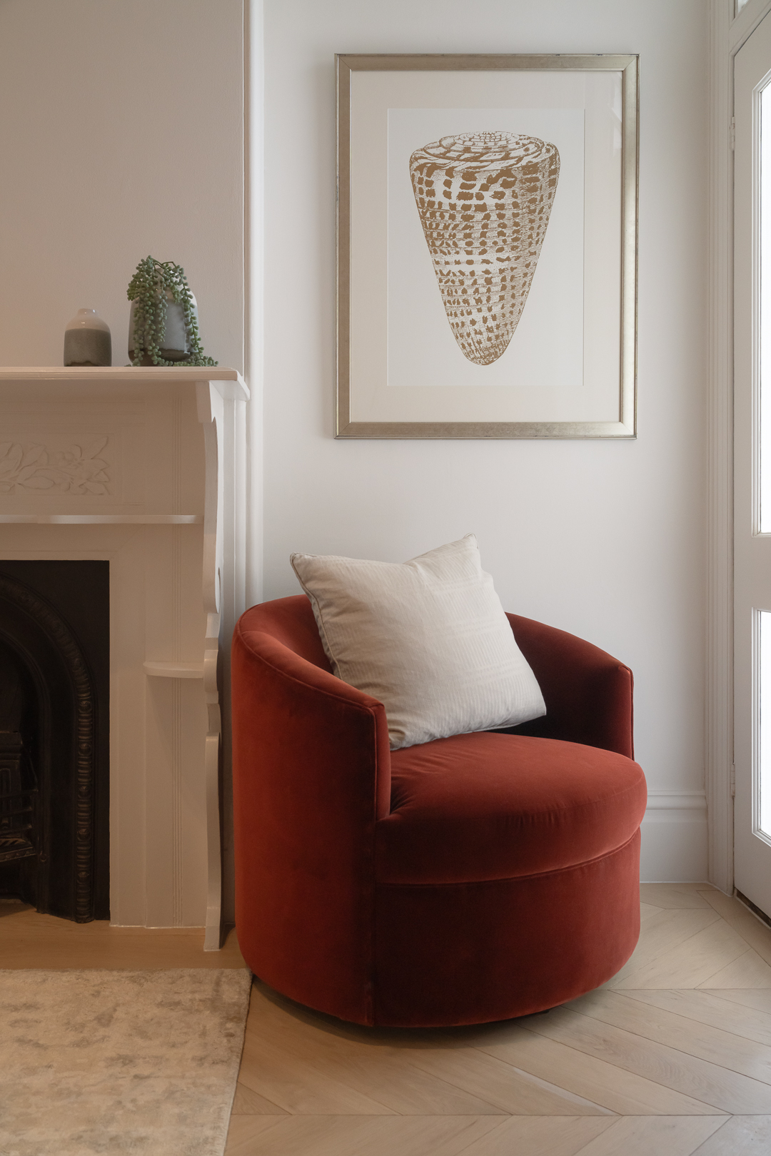 Interior view of living room with rust-colored velvet barrel chair, white pillow, fireplace, framed art, and herringbone wood floor.