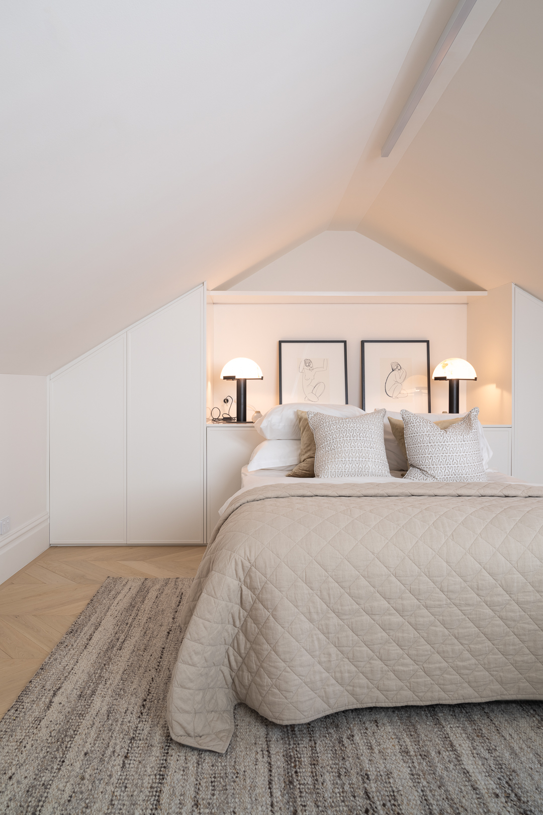 Bedroom interior with a bed covered in a beige quilted comforter, two lamps on bedside tables, and a woven rug on a light wood floor.