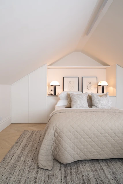 Bedroom interior with a bed covered in a beige quilted comforter, two lamps on bedside tables, and a woven rug on a light wood floor.