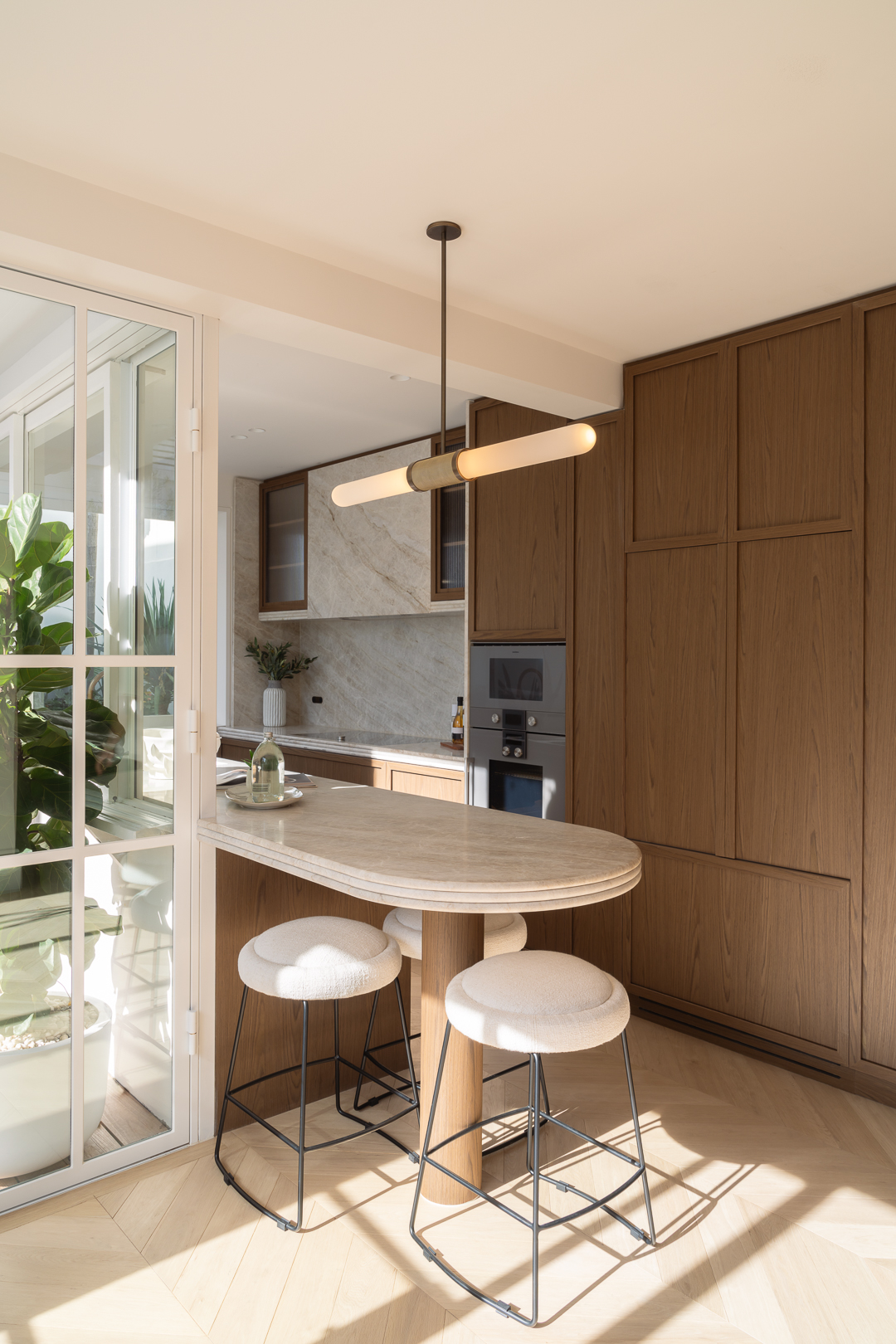 A modern kitchen interior features a marble island with bar stools, wood cabinets, stainless steel appliances, and a view through glass doors.