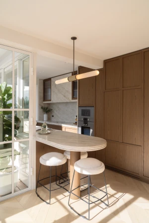 A modern kitchen interior features a marble island with bar stools, wood cabinets, stainless steel appliances, and a view through glass doors.