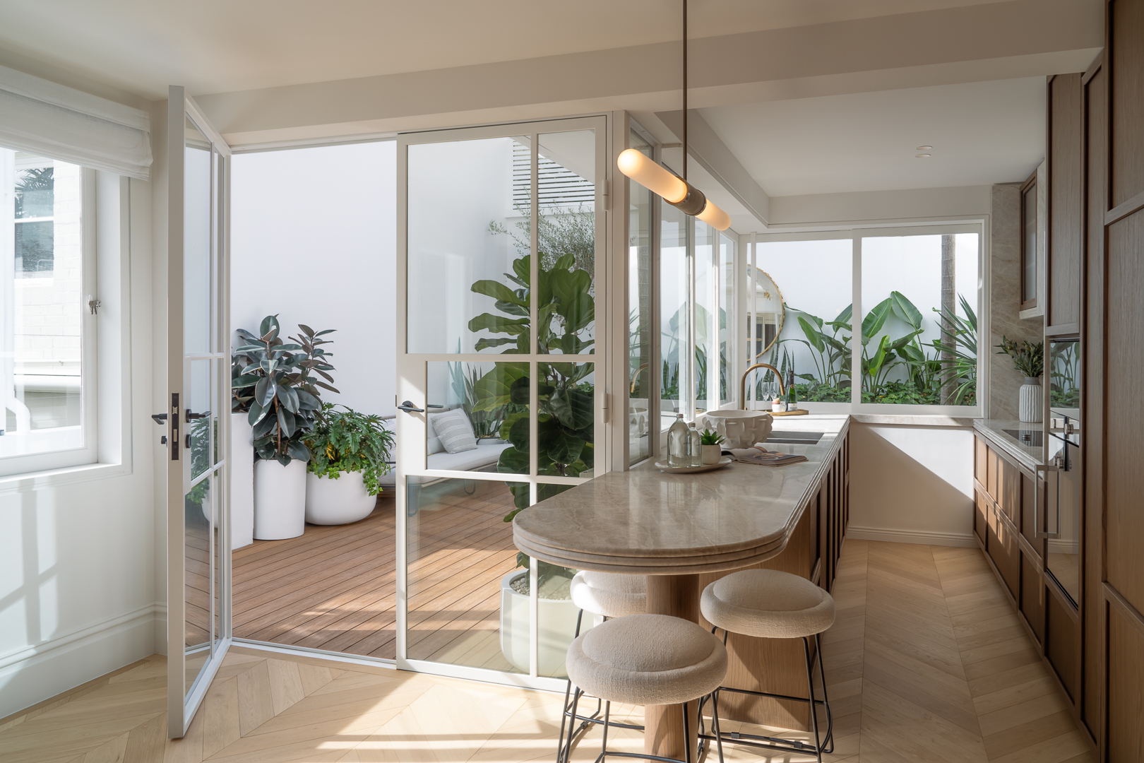 Modern kitchen with marble island and bar stools opens onto an outdoor courtyard filled with green plants and bright natural light.