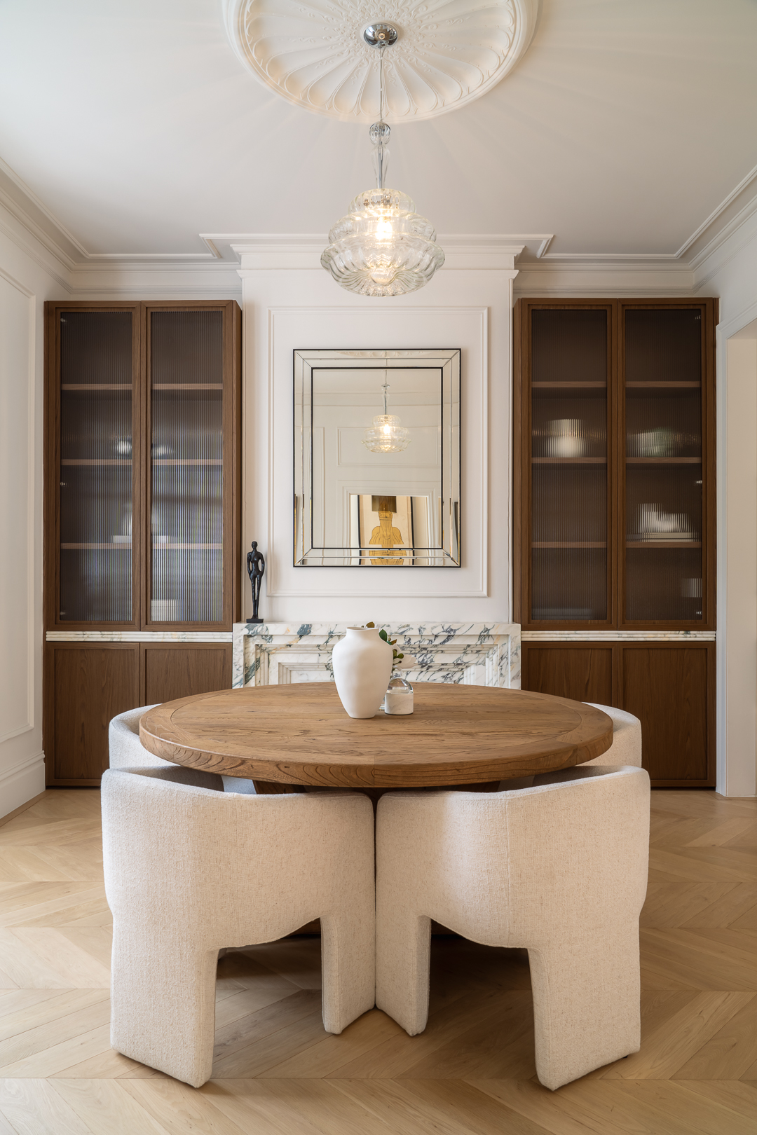 Interior of a dining area featuring a round wooden table surrounded by modern white chairs, with built-in dark wood shelving.