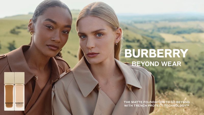 Cosmetics advertisement featuring two women with fair skin tones wearing tan trench coats, product containers, and a green and brown hillside backdrop.
